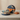A round gift box with a blue and gold floral lid and orange ribbon, filled with assorted chocolates, sits open next to a matching closed box and a small wrapped present on a beige surface.